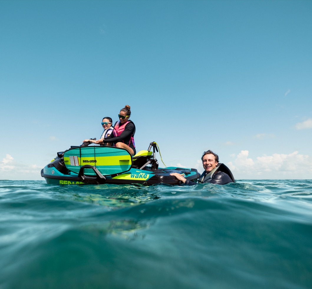 Zwei Frauen sitzen auf einem Sea-Doo Wake Pro, während sich ein Mann im Wasser an der ST3-Plattform festhält